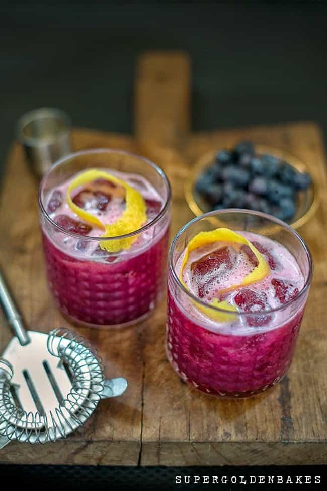 Two glasses of blueberry gin sour cocktails garnished with lemon peel