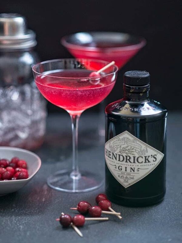 A bottle of Hendrick’s Gin sits next to a martini glass filled with a bright pink Red Queen cocktail, garnished with berries. Another gin cranberry St Germain drink, a cocktail shaker, and a dish of red berries are in the background.