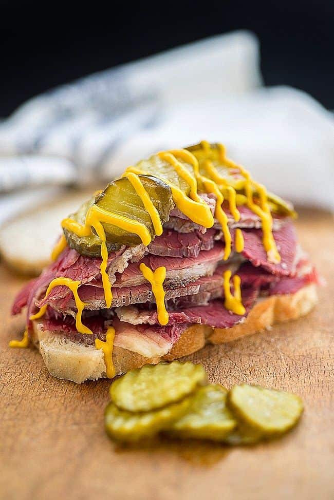 Close up on an open salt beef sandwich topped with pickles and yellow mustard
