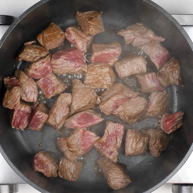 Chunks of beef for a hearty beef stifado are seared in a black frying pan on the stovetop, some still pink in the center and others browned at the edges, with steam rising from this classic Greek beef stew.