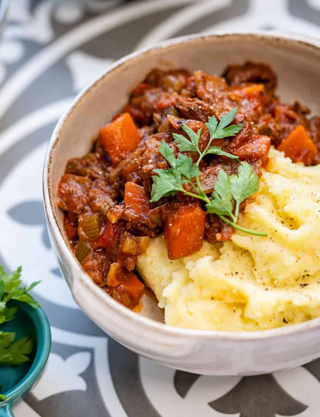 A bowl of mashed potatoes topped with a hearty Greek beef stew, garnished with fresh parsley, sits on a decorative plate.