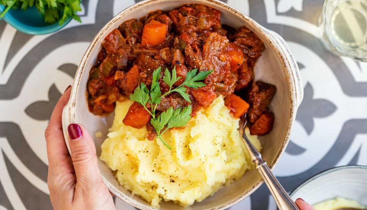 A bowl of mashed potatoes topped with beef stifado and garnished with parsley, held by a person with red nail polish. A fork rests in the bowl on a patterned table surface.