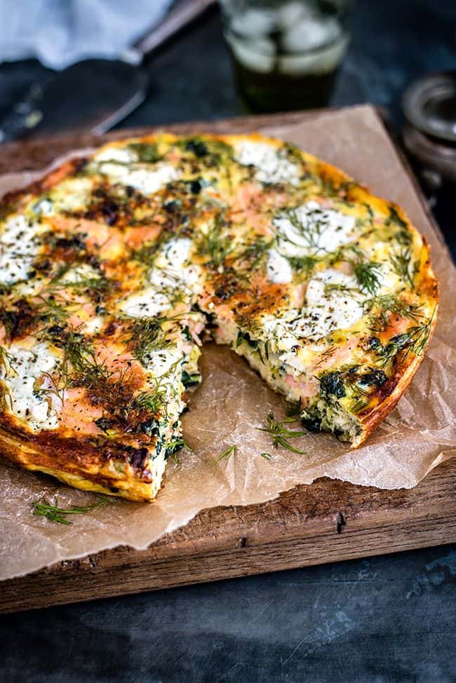 A savory cottage cheese and smoked salmon frittata with a slice missing, featuring spinach and herb toppings, lies on parchment paper over a wooden board.
