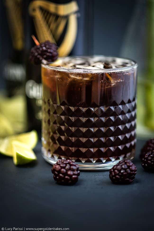A glass of Black Magic cocktail with ice sits on a table, garnished with a blackberry. Fresh blackberries and lime wedges are scattered nearby, with a blurred dark bottle in the background.