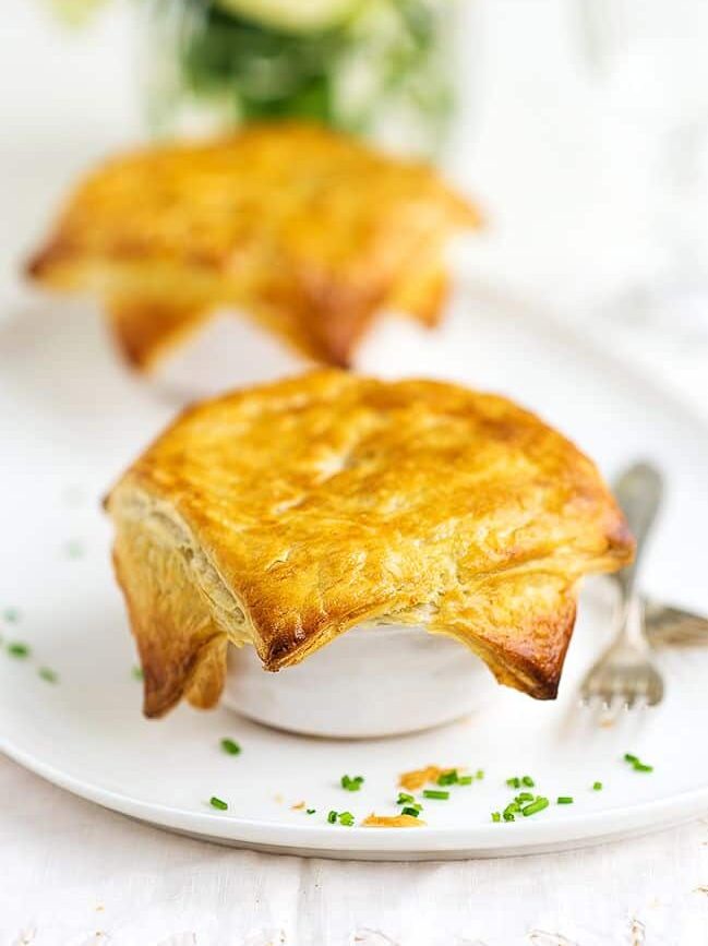 Two golden-brown lobster pot pies made with puff pastry sit on a white plate, sprinkled with green herbs, and a fork placed beside one of the pies.
