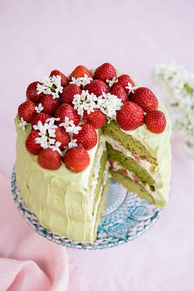 A tall, round Matcha strawberry layer cake with pale green frosting is topped with fresh strawberries and small white flowers on a decorative stand. A slice has been cut, revealing vibrant green cake layers inside.
