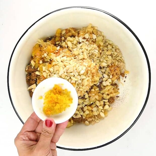 A hand with red-painted nails holds a small dish of orange zest over a mixing bowl filled with chopped nuts, dried fruit, and sugar—essential ingredients for a classic Christmas pudding—on a white background.