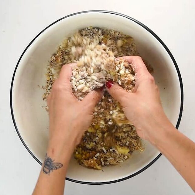 Two hands mix granola ingredients in a white bowl, the left wrist adorned with a bee tattoo and red-painted nails. Oats, seeds, dried fruit—and hints reminiscent of christmas pudding—add festive flair to the wholesome blend.