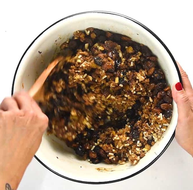 A person stirs a bowl of chunky, sticky christmas pudding fruit and nut mixture with a wooden spoon. Only their hands are visible—one holding the bowl, the other stirring the festive blend.