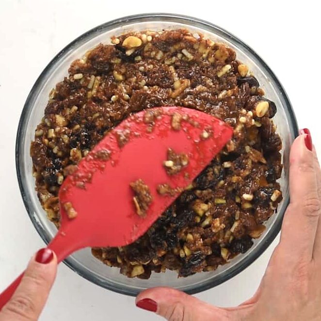 A person with red nail polish uses a red spatula to mix a thick, chunky christmas pudding batter with nuts and raisins in a glass bowl on a white surface.