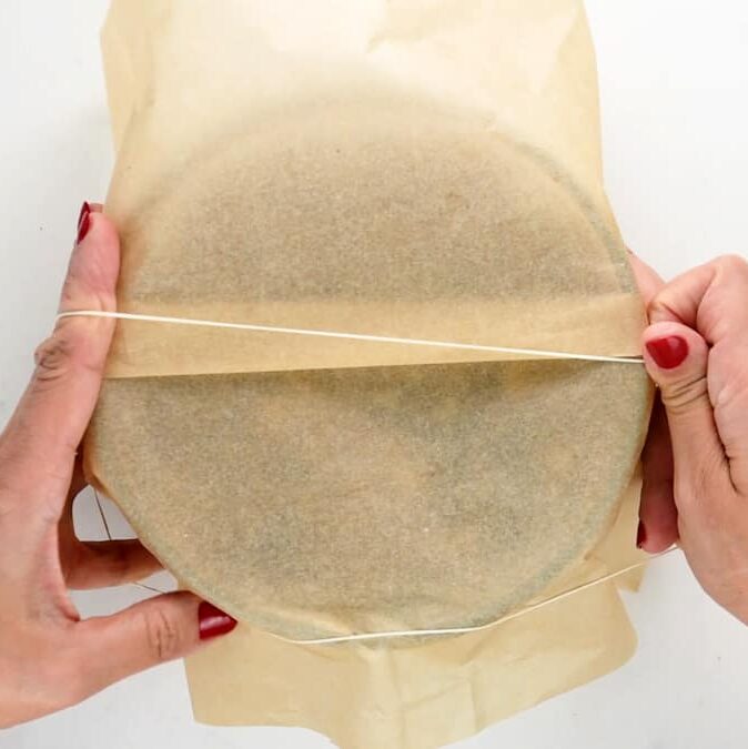 Two hands with red nail polish stretch a rubber band around a round baking pan covered with parchment paper, preparing it for a classic Christmas pudding.