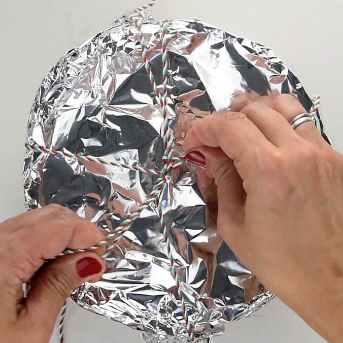Hands tying a red, white, and green striped string over a bowl covered with crinkled aluminum foil—securing a Christmas pudding for steaming. The person has red nail polish and is wearing a ring.