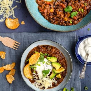 Bowl of lentil and bean chili topped with sour cream, avocados, grated cheese and Popchips