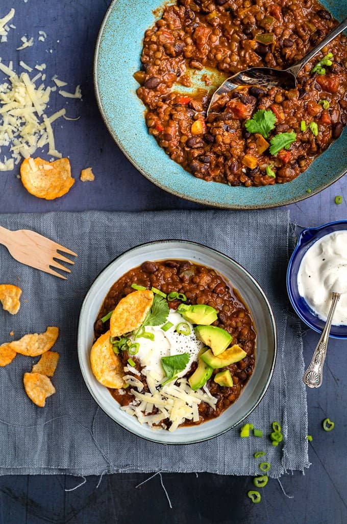 Bowl of lentil and bean chili topped with sour cream, avocados, grated cheese and Popchips