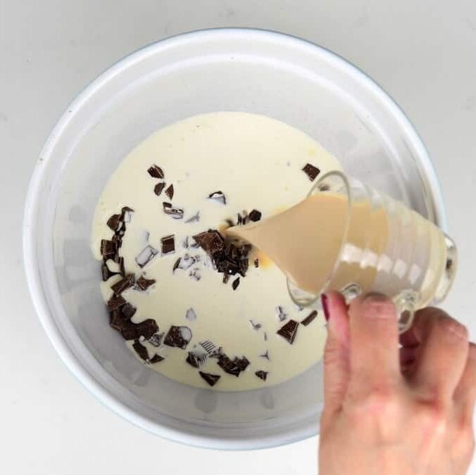 A hand pours a creamy liquid from a glass measuring jug into a bowl containing cream and chopped chocolate pieces, starting this easy Baileys chocolate mousse recipe.