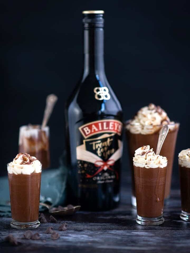A bottle of Baileys Irish Cream stands behind five small glasses of easy Baileys Chocolate Mousse, each topped with whipped cream and chocolate shavings, against a dark background.