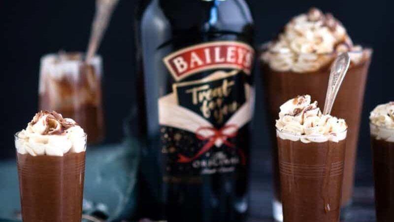 Glasses of chocolate mousse topped with whipped cream and chocolate shavings are arranged around a bottle of Baileys Irish Cream—an easy Baileys chocolate mousse perfect for pudding lovers.