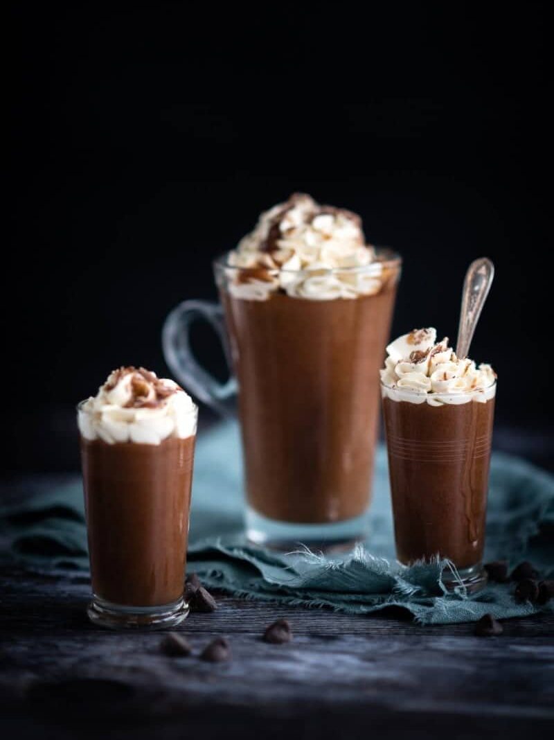 Three glass mugs filled with rich hot chocolate, each topped with swirls of whipped cream and a sprinkle of chocolate shavings, evoke the indulgence of Baileys Chocolate Mousse. A blue cloth and scattered chocolate chips complete the cosy scene.