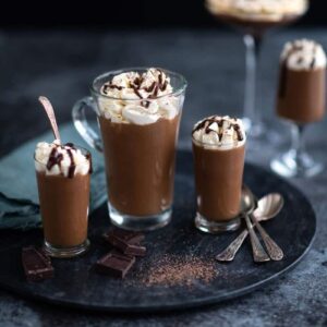 Four glasses of creamy Baileys chocolate mousse topped with whipped cream and chocolate drizzle are arranged on a dark round tray, accompanied by silver spoons and chocolate pieces. The background is dark and blurred.