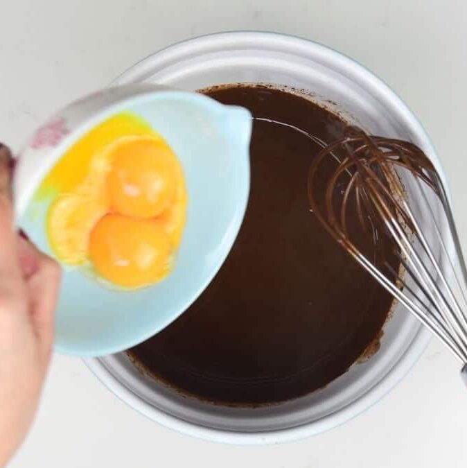 A hand holds a small bowl with three cracked eggs, pouring them into a larger bowl filled with a dark liquid mixture for easy Baileys Chocolate Mousse. A metal whisk rests inside the larger bowl on a white surface.