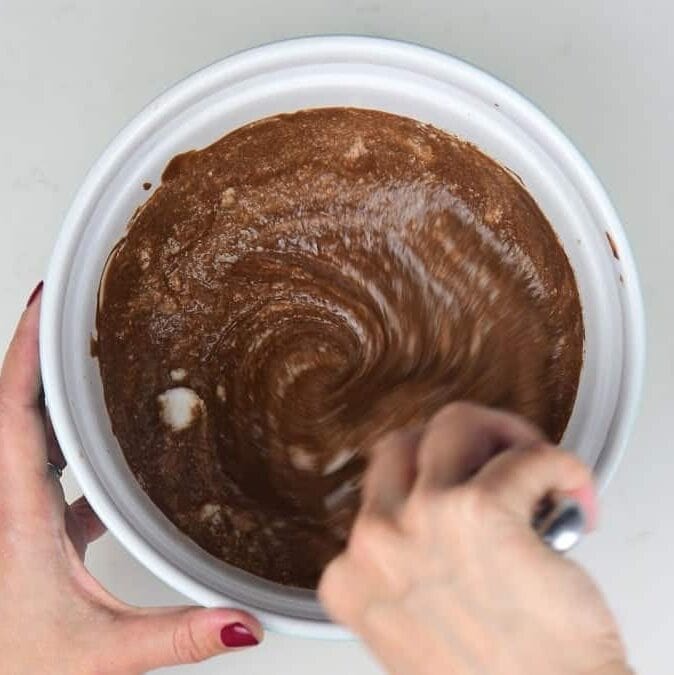 Hands mixing chocolate batter in a white bowl with a whisk, photographed from above on a white surface. The thick mixture is being stirred, showing an early step in a Baileys Chocolate Mousse recipe.