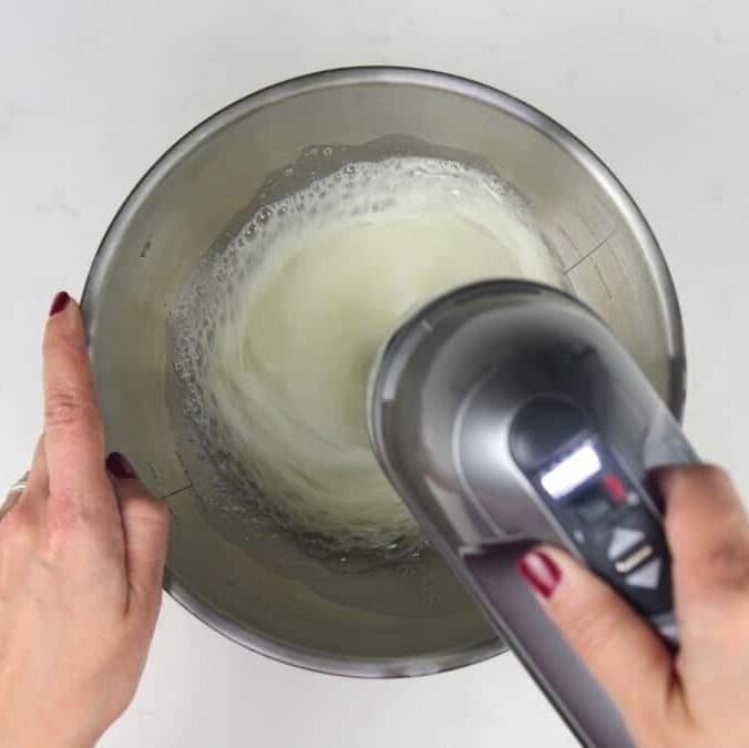 A person uses a hand mixer to whisk egg whites in a stainless steel mixing bowl, preparing for an easy Baileys chocolate mousse. The person's fingernails are painted red.