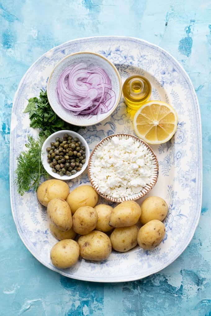 Ingredients for Greek Potato Salad on a platter: baby potatoes, red onion, feta, capers, herbs, lemon, olive oil