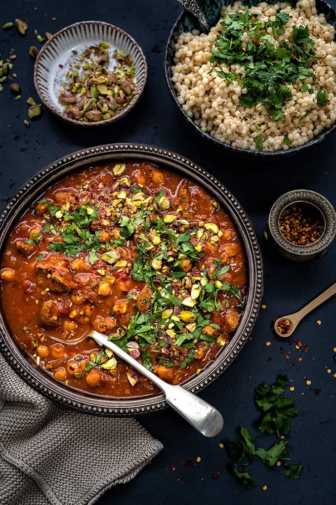 Overhead shot of lamb tagine in a rustic metal bowl with giant couscous on the side on dark background