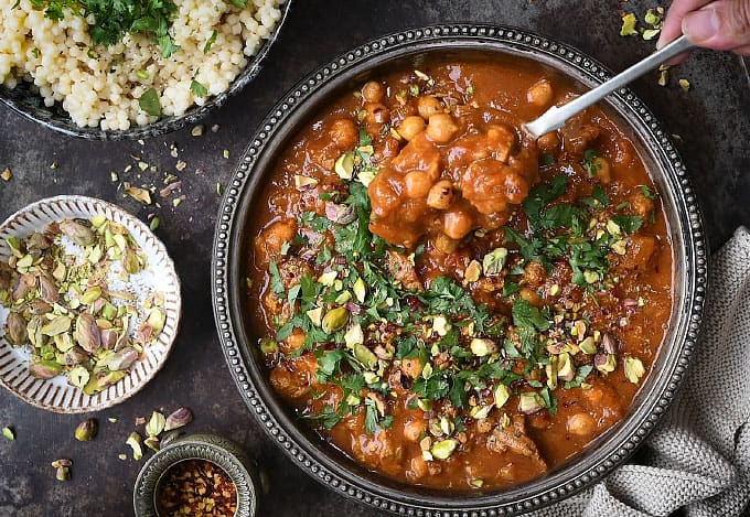 Lamb tagine in a rustic metal bowl with giant couscous on the side on dark background