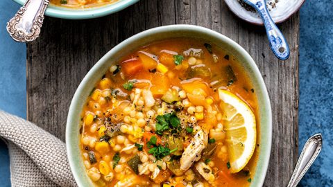 Two bowls of Hearty Chicken Vegetable Soup with pearl barley, garnished with lemon wedges and fresh herbs, sit on a wooden surface beside a spoon, pepper dish, beige napkin, and a bottle of olive oil.