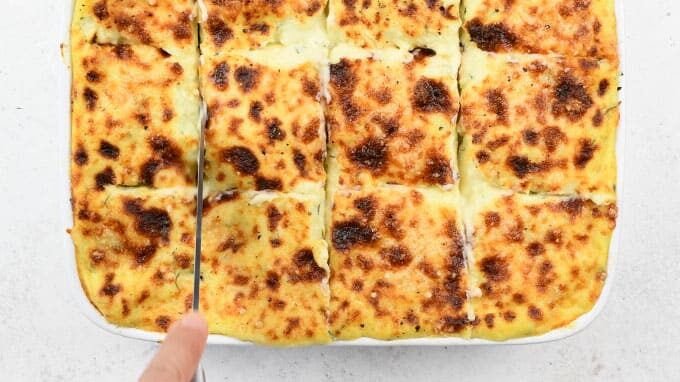 A hand is cutting a baked dish, likely salmon lasagna, into nine square portions. The top layer is golden brown and slightly charred. The dish sits in a white rectangular baking pan on a white surface.