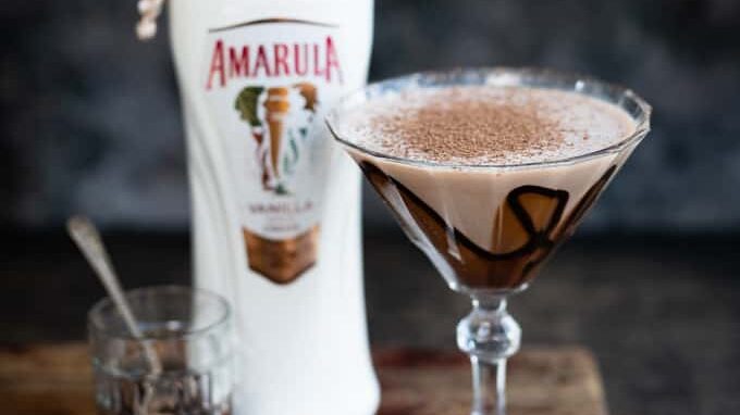 A Chocolate Martini fills a martini glass, creamy and drizzled with chocolate syrup, topped with cocoa powder. It sits beside a bottle of Amarula Vanilla and a small glass with a spoon on a wooden surface.