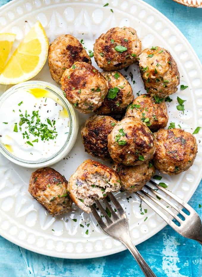 A white plate with browned Greek turkey meatballs garnished with chopped parsley, served with a small bowl of authentic tzatziki sauce topped with herbs and lemon wedges. Two forks rest on the plate over a blue background.