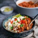 A bowl of slow cooker chili served over rice, topped with sour cream, grated cheese, cilantro, and sliced red chili peppers, with a lime wedge on the side. Another bowl of chili con carne is blurred in the background.