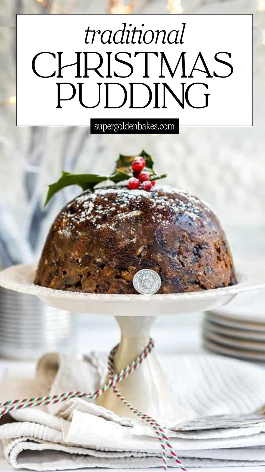 A classic Christmas pudding topped with holly and berries sits on a cake stand, dusted with powdered sugar. Plates are stacked in the background, and festive string decorates the base—perfect for a traditional holiday celebration.
