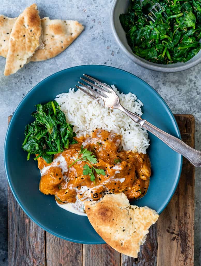 Chicken curry served in a blue bowl with rice, naan bread and steamed spinach
