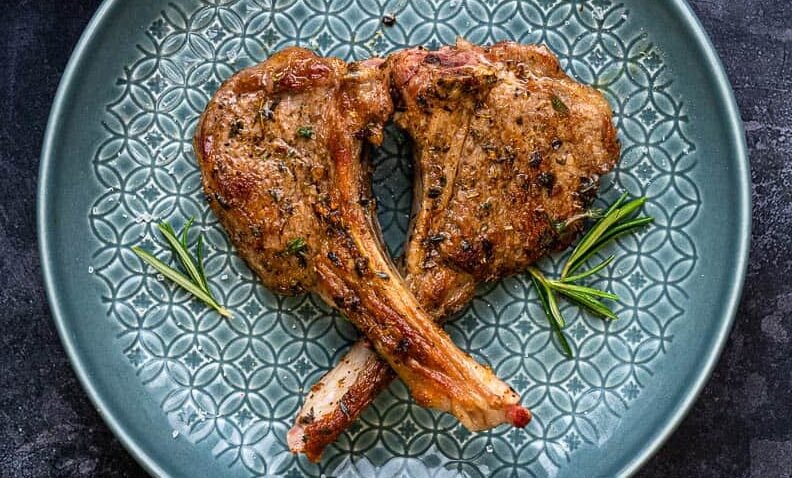 Two succulent lamb chops arranged in a heart shape on a textured blue plate, garnished with fresh rosemary sprigs.