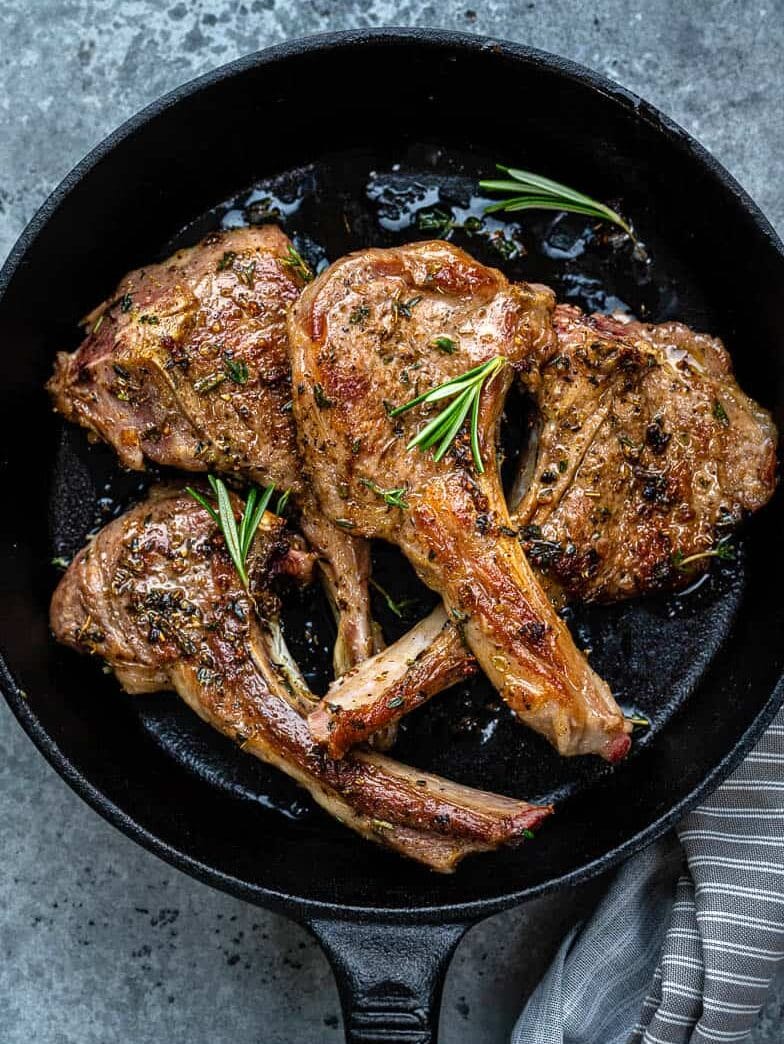 Three cooked lamb chops with herbs and seasoning are arranged in a black cast iron skillet, garnished with fresh rosemary sprigs. A striped cloth napkin is partially visible at the skillet’s edge, completing the inviting display of savory lamb chops.