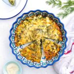 A crustless quiche with spinach and feta sits in a decorative blue pie dish, garnished with fresh dill. Two slices are cut and slightly separated. A cup of grated cheese and a white tea towel are nearby on a white surface.