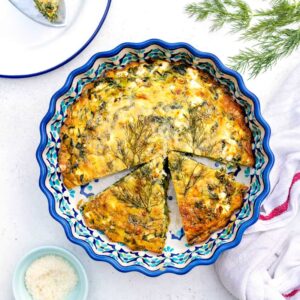 A crustless quiche with spinach and feta sits in a decorative blue pie dish, garnished with fresh dill. Two slices are cut and slightly separated. A cup of grated cheese and a white tea towel are nearby on a white surface.