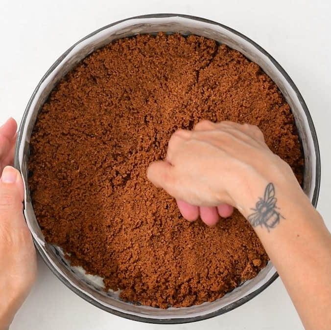 A person presses a crumbly Biscoff no bake cheesecake crust into a round baking pan. One hand holds the pan while the other presses down; a bee tattoo is visible on their wrist. The background features a white surface.