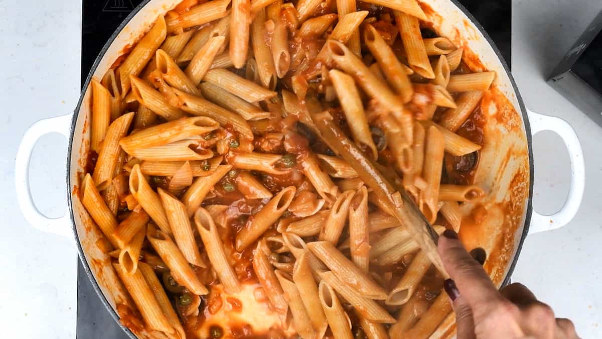 Tossing cooked penne in tomato olive and caper sauce