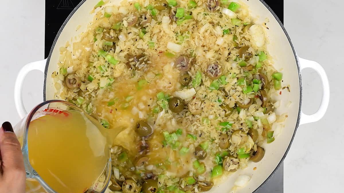 rice cooking with onions, pepper and olives in pan with chicken stock being poured in