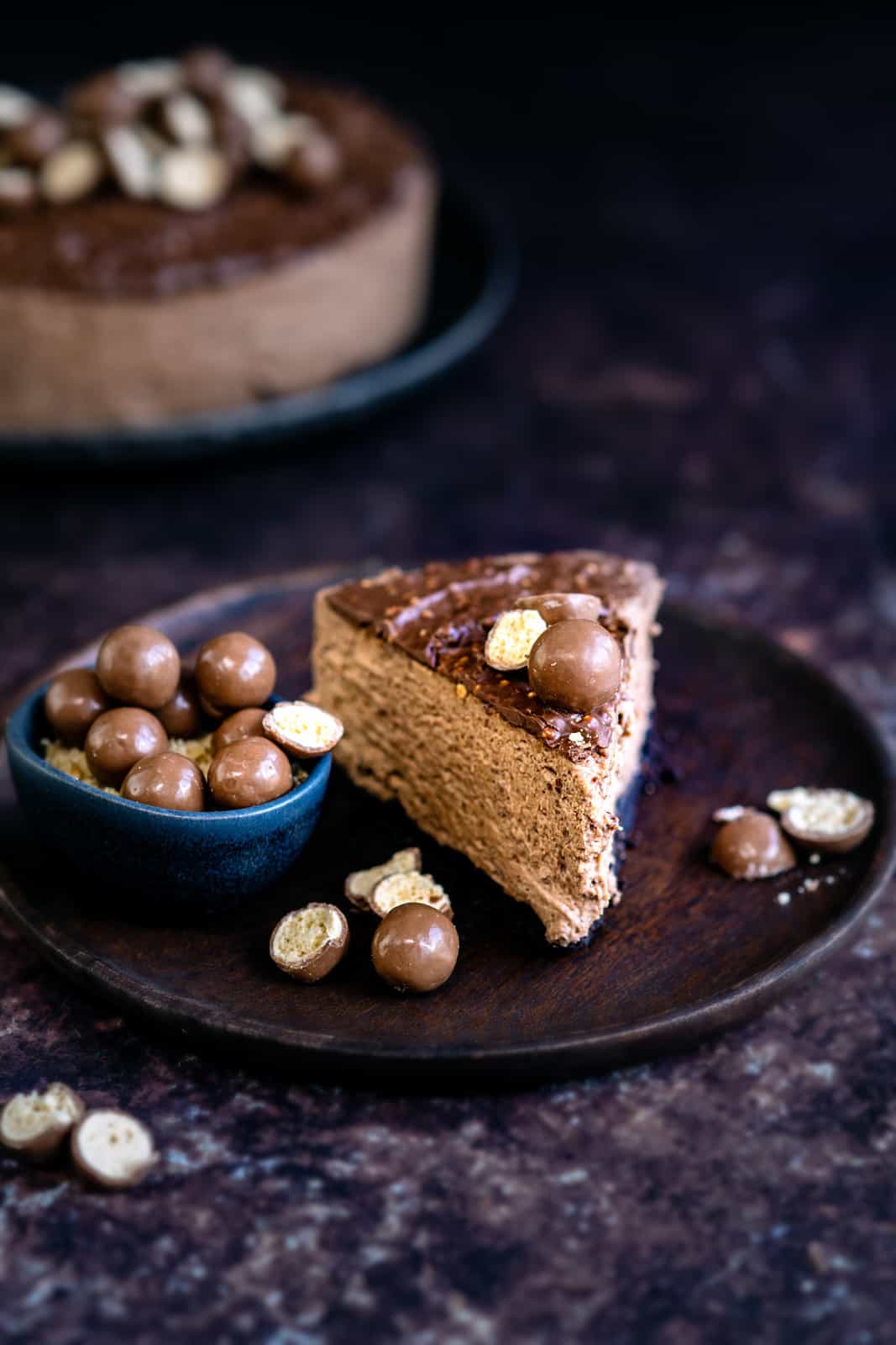 Slice of Malteser cheesecake on wood plate with Malteses on the side