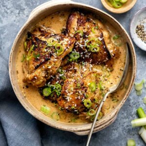 A bowl of creamy, golden-brown slow cooker chicken thighs garnished with sliced green onions and grated cheese, served with a spoon on a gray surface, next to a small dish of chopped green onions and a bowl of pepper.
