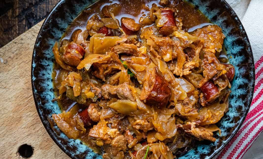 A bowl of hearty Bigos, or Polish Hunter's stew, featuring shredded cabbage, chunks of sausage, and pieces of pork in a rich brown sauce, served on a wooden surface with a striped cloth partially visible.