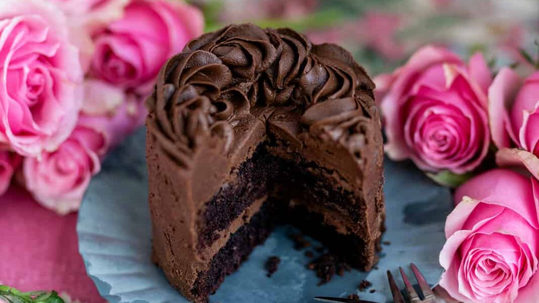 A mini chocolate cake for 2 with rich frosting and floral decorations sits on a blue paper liner, surrounded by pink roses. A slice has been removed, revealing its moist, dark interior. A fork lies nearby.
