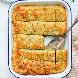 A rectangular baking dish with golden-brown, sesame-topped Greek spanakopita cut into squares. A serving spatula rests in the dish, with one spinach pie slice missing. Fresh dill and a plate with a serving are nearby.