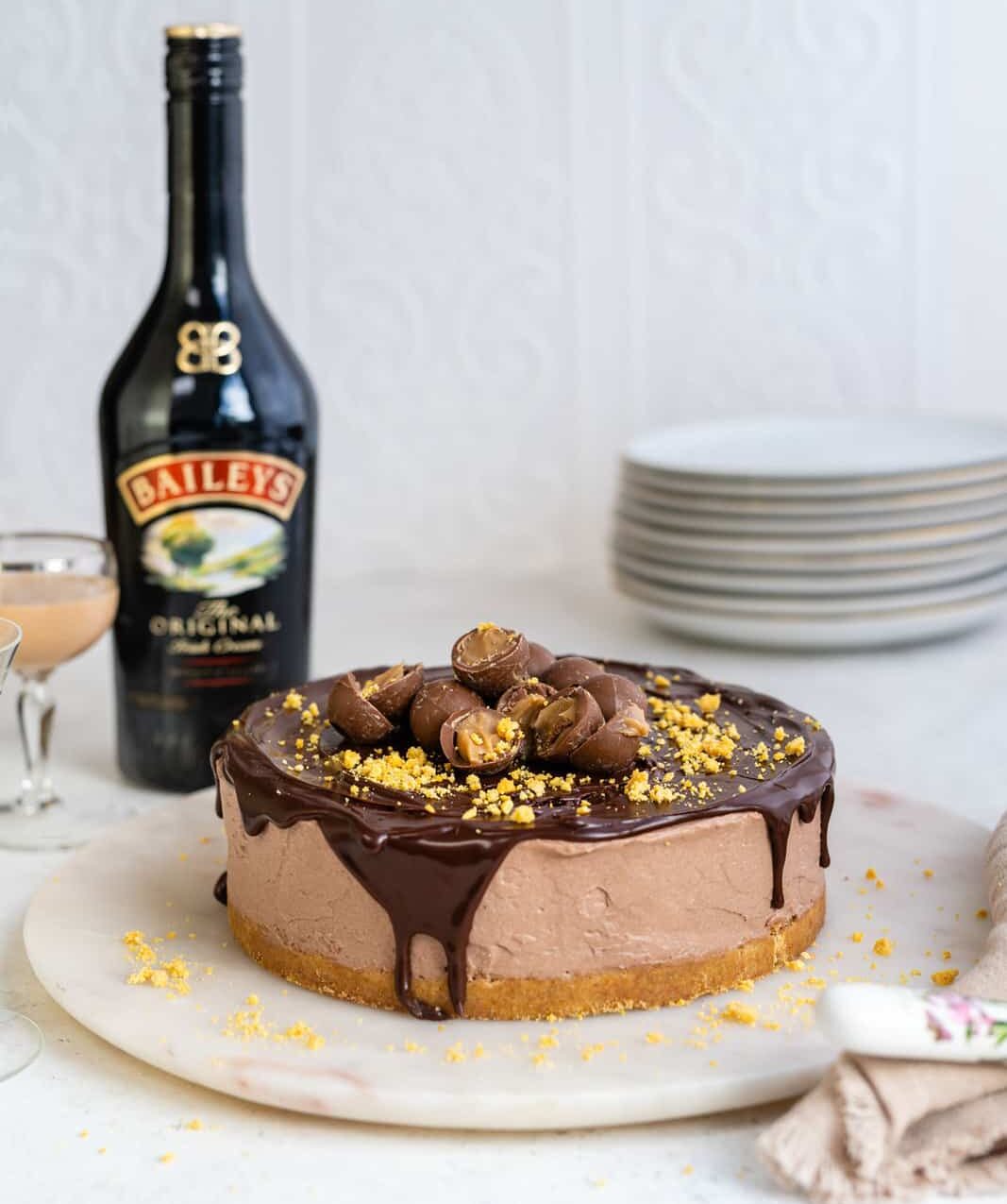 A no bake Baileys Cheesecake topped with chocolate glaze, crushed yellow sprinkles, and chocolate truffles sits on a marble board. Behind it, there’s a Baileys Irish Cream bottle, a glass of liqueur, and stacked plates.