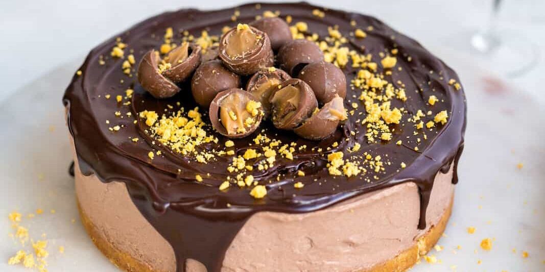 A no bake Baileys Cheesecake topped with chocolate drizzle, halved chocolate candies, and crushed crumbs sits on a marble surface. Plates are stacked in the background with a glass of dessert drink nearby.